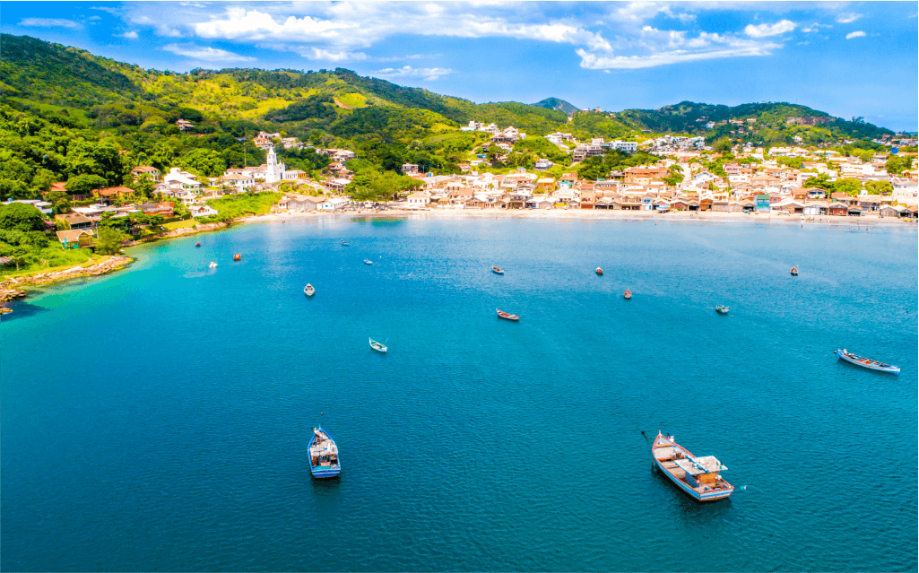 Imagem do mar bem azul cercado por montes verdes com casa. O mar de Garopaba está repleto de barcos.