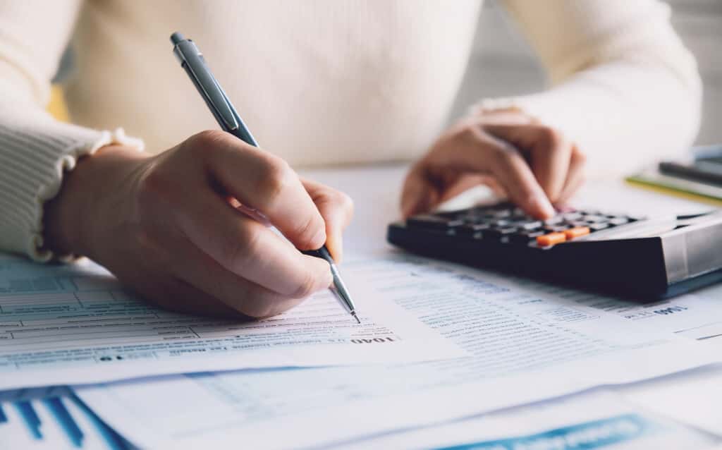 Imagem que mostra uma as maõs de uma mulher apoiadas em cima de uma mesa enquanto ela escreve com a mão direita e usa uma calculadora com a mão esquerda para pesquisar o informe de rendimentos