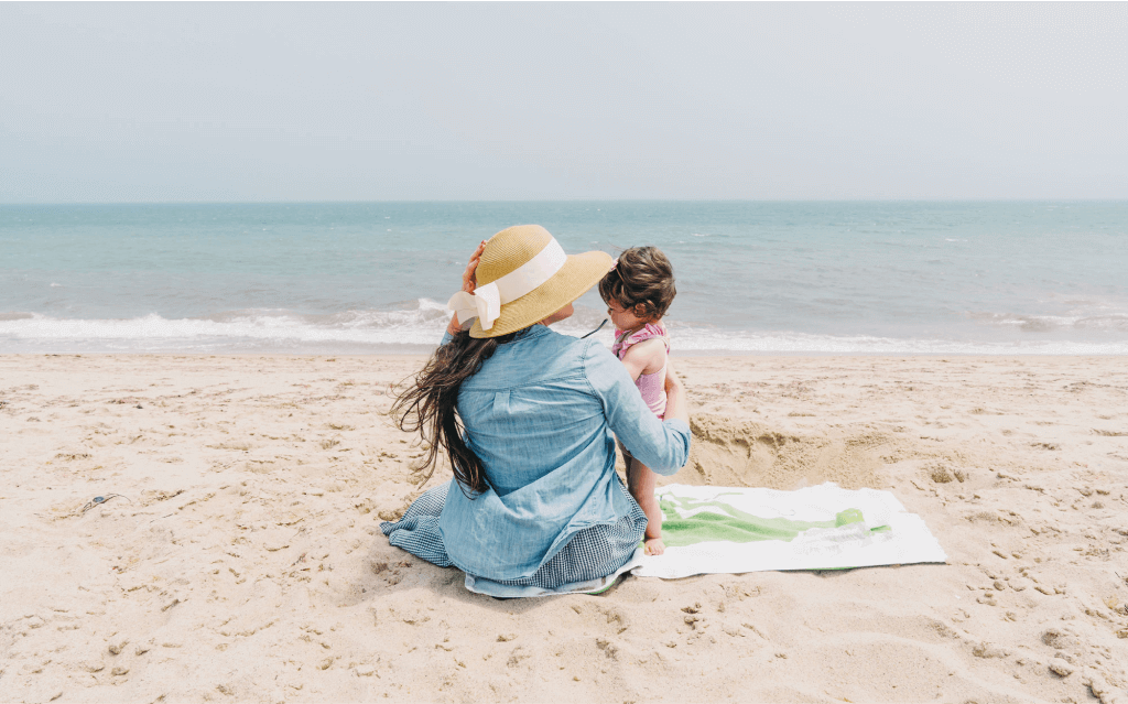 mulher sentada sobre uma canga na areia de uma praia vestida com uma blusa de mangas longas e um chapéu de palha com um laço. Ela está segurando um bebê, simbolizando viagens em junho