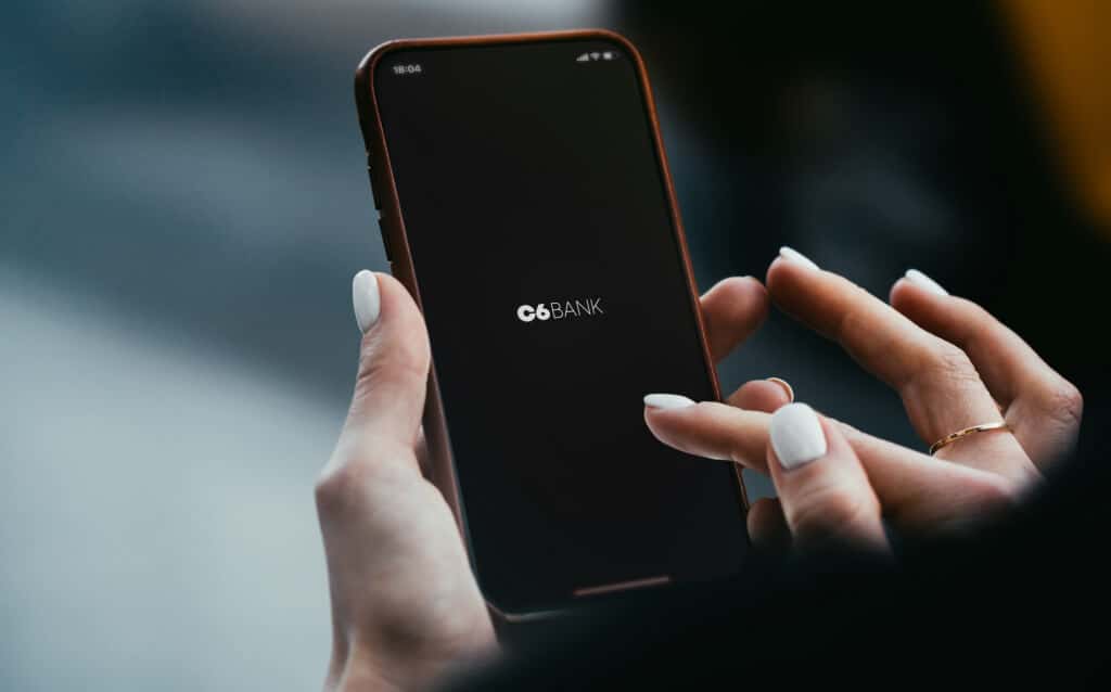 Mão segurando um celular escrito C6 Bank na tela para ilustrar o funcionamento do câmbio programado no app
