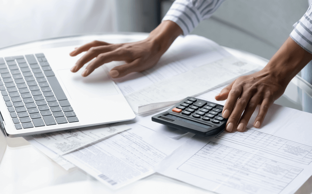 Mãos de uma pessoa negra digitando em uma calculadora com a mão direita e em um notebook com a mão esquerda. Na mesa onde os objetos estão apoiados, tem algumas folhas de papel