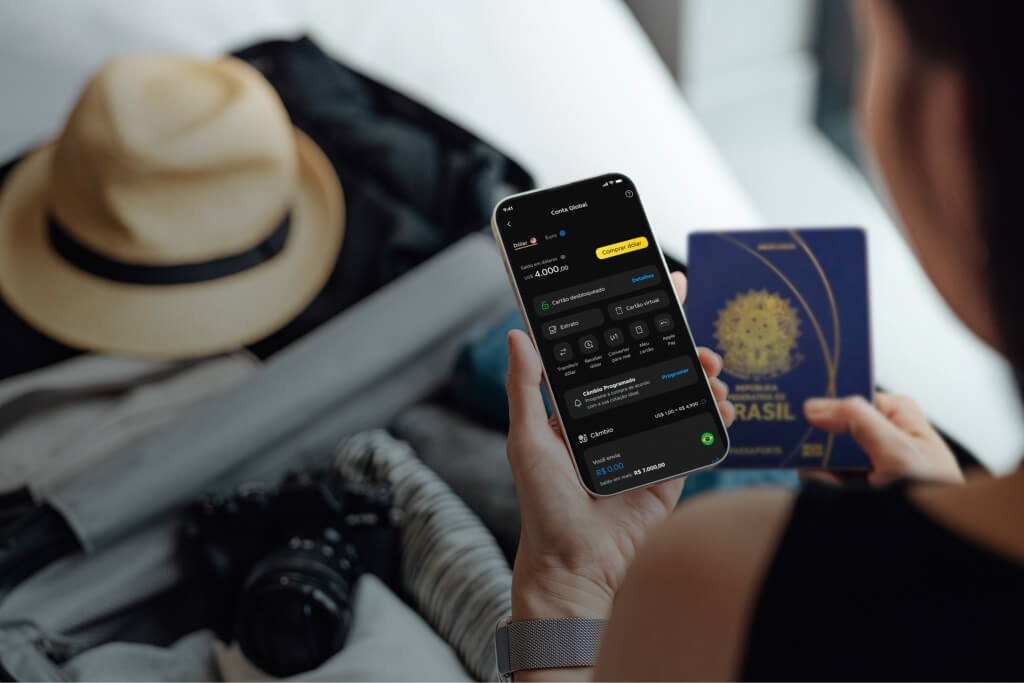 Mulher de costas segurando um celular com a mão direita que mostra o aplicativo do C6 Bank aberto e um saldo de 4 mil reais, e um passaporte na mão esquerda. À sua frente tem uma mala com um chapéu em cima