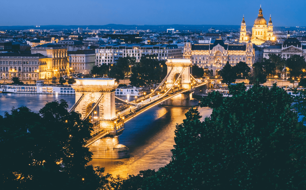 Uma grande ponte iluminada passa por cima de um rio durante a noite na cidade de Budapeste, cercada por construções e prédios.