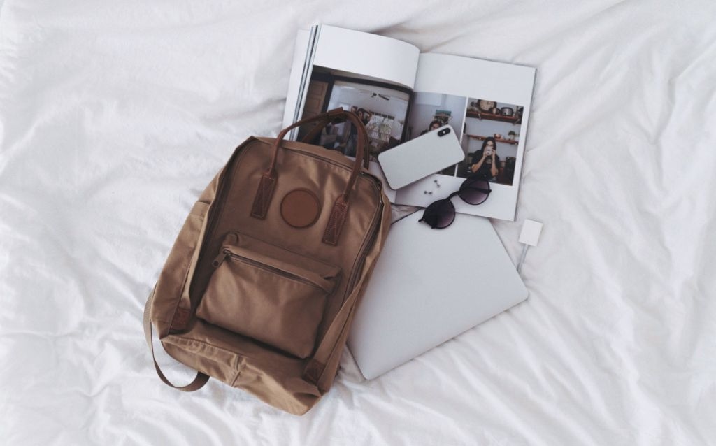 viagem feriado prolongado itens de viagem sobre a cama óculos de sol, celular, mochila marrom de tecido, revista , notebook