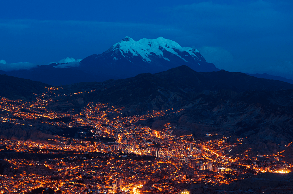 Vista de cima da cidade de La Paz, com a luzes da cidade na frente e montanhas com neve atrás.