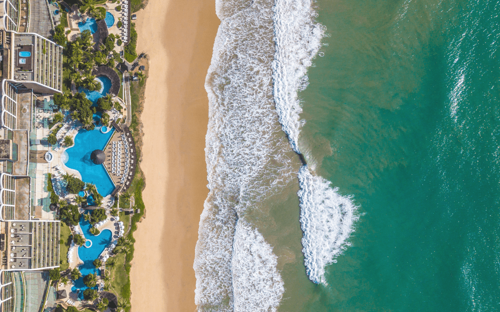 Vista de cima da praia. É possível ver a areia, o mar e a área de lazer de um prédio que é pé na areia.