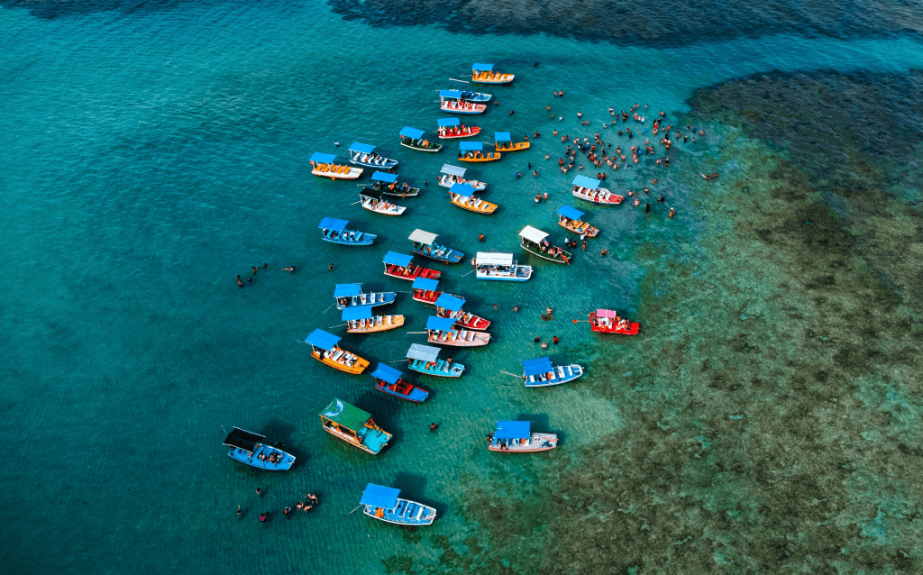 Vista de cima do mar transparente com tonalidade esverdeada e diversos barcos coloridos boiando na água.