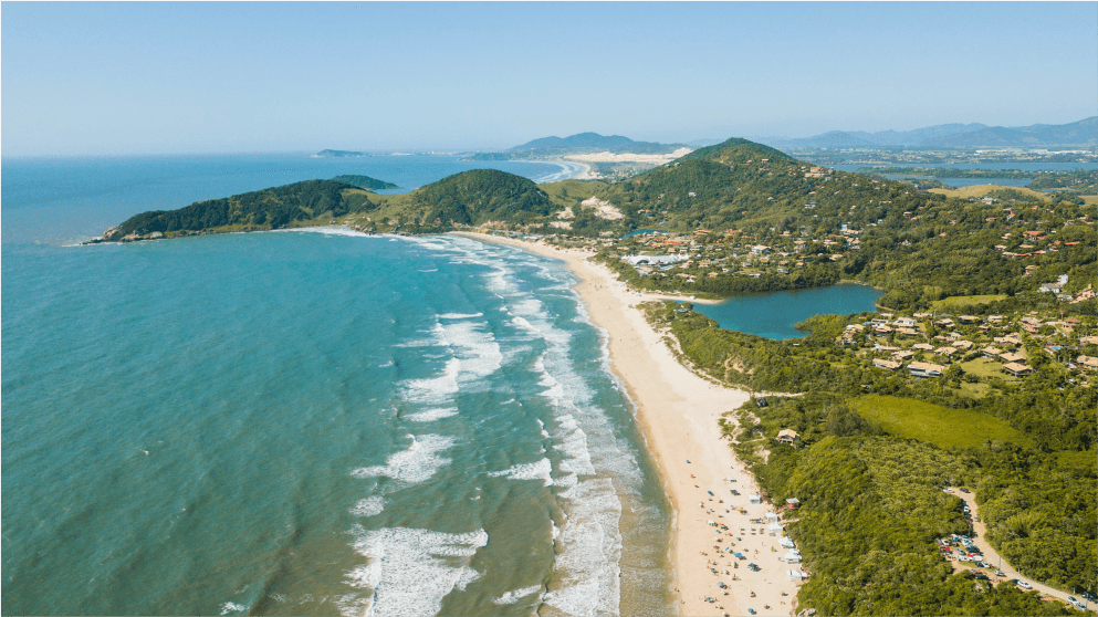 Vista panorâmica da Praia do Rosa em Florianópolis. É possível ver uma lagoa, o mar e montes verdes da região.