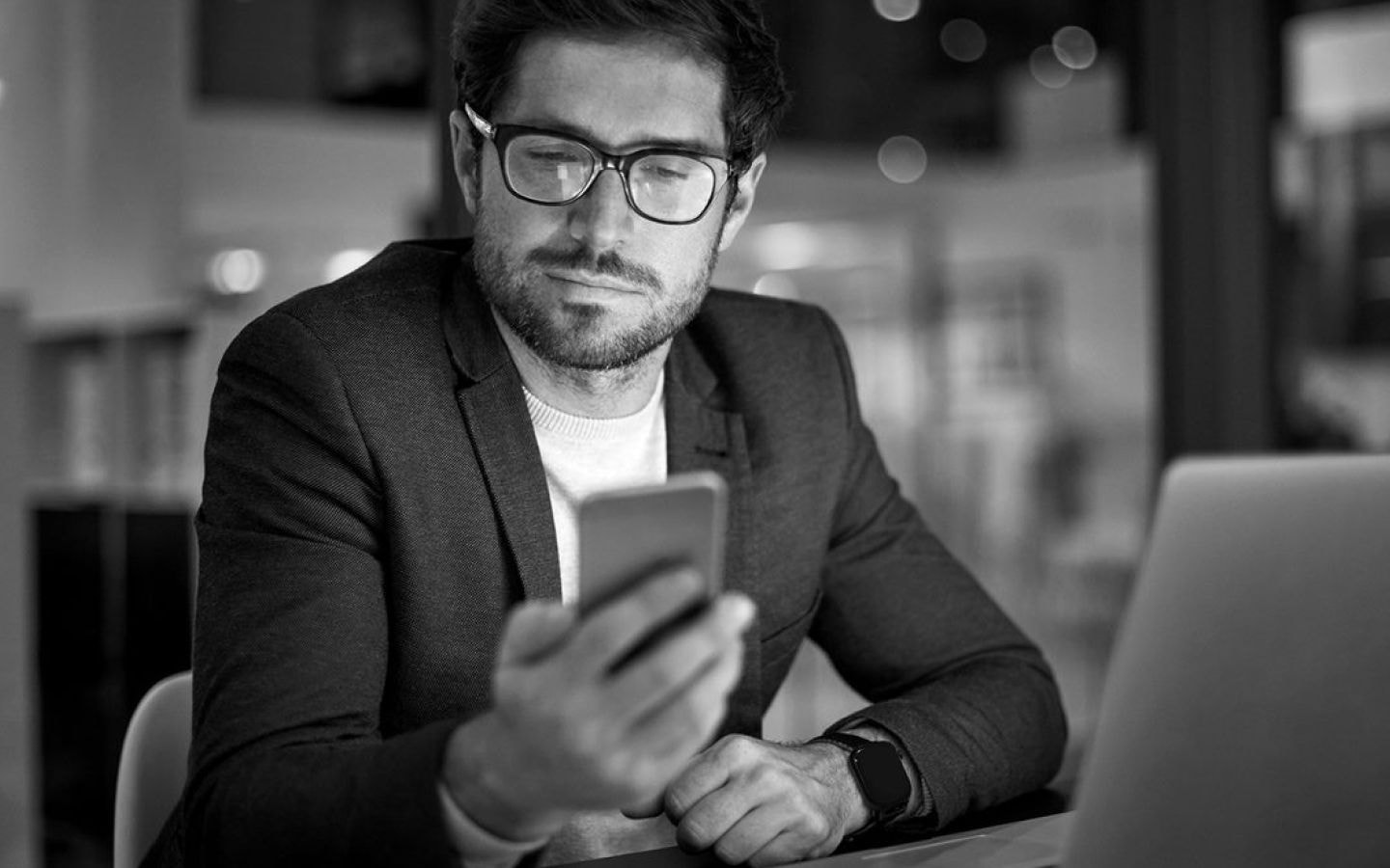 Foto de um homem branco, de cabelos lisos curtos. Ele segura um celular em sua mão direita. A foto está em preto e branco. Ele usa camiseta, blazer e óculos e está sentado em frente à um notebook.