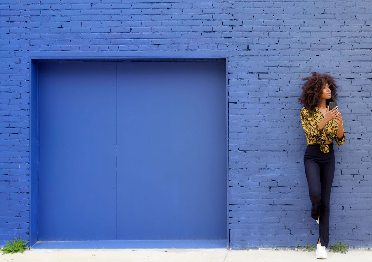 Foto de uma mulher negra, de cabelos crespos, castanhos e longos. Ela segura um celular em sua mão direita. Usa blusa amarela com listras pretas e calça azul escura. Está de pé em frente à uma parede de tijolos pintada de azul claro.
