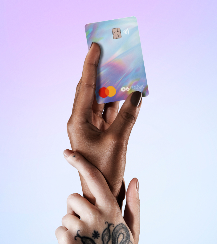 Uma mão branca com tatuagem entrelaçada com uma mão negra segurando o Cartão de Crédito C6 Rainbow