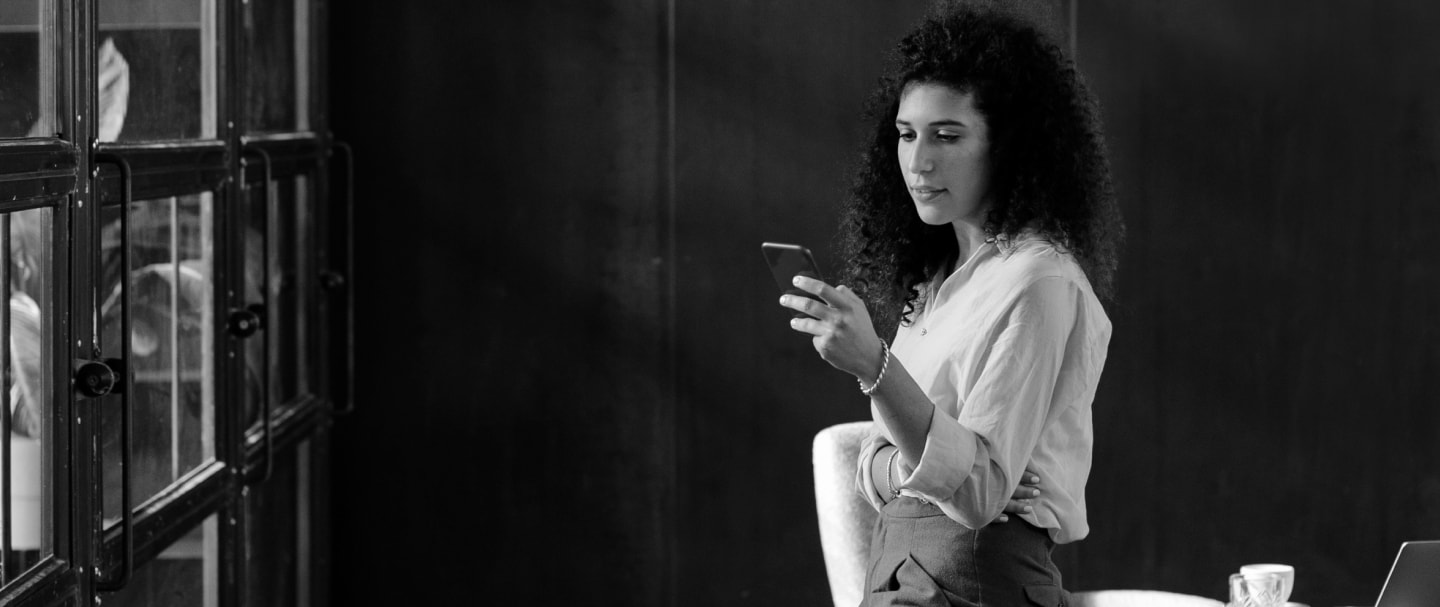 Modelo feminina de cabelos cacheados de perfil em foto preto e branco, na mão esquerda ela segura um celular.