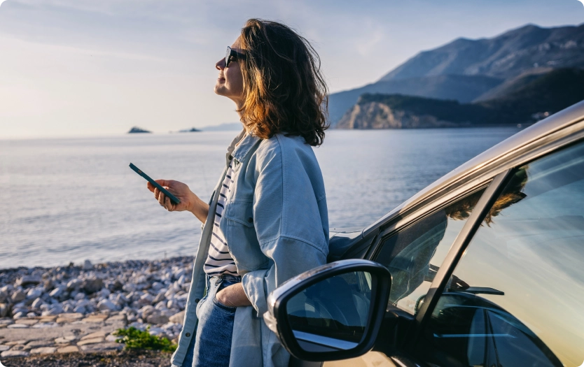 Fotografia de mulher de cabelo curto, ondulado e castanho, usando óculos escuro, apoiada na lateral de um carro, segurando um celular com a mão direita e observando a paisagem de lago e montanhas.