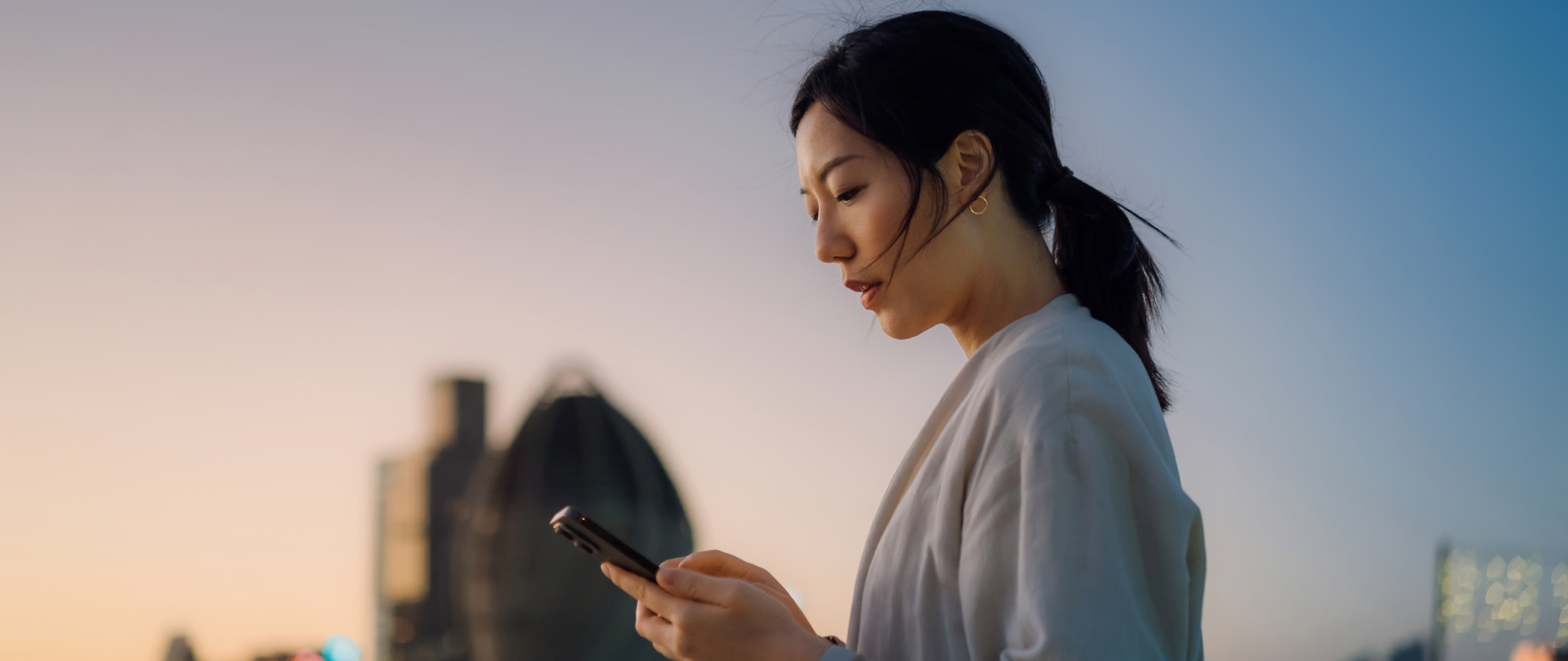 Câmbio e comex Mulher usa celular ao ar livre, com cidade desfocada ao fundo no entardecer.