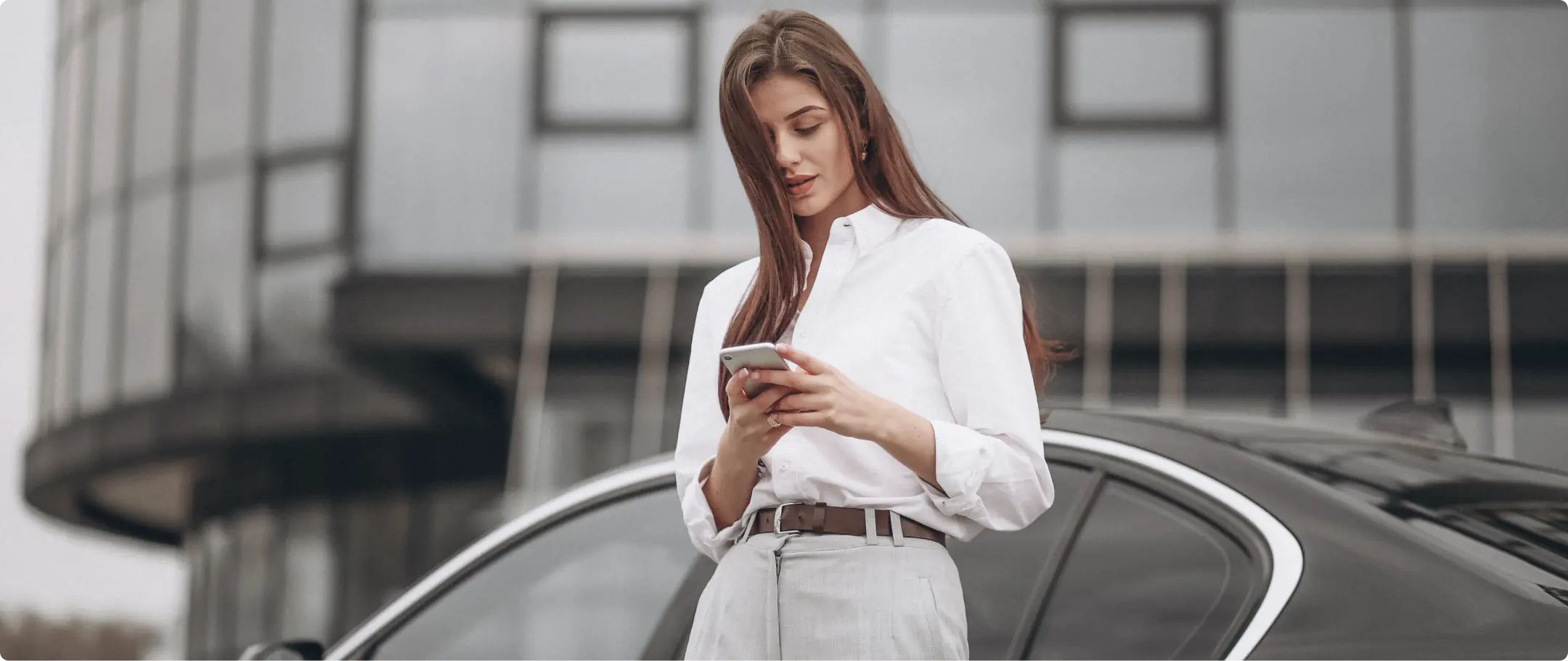 Mulher Car Equity Mulher com cabelo castanho, vestindo camisa social branca para dentro da calça, segurando um celular e encostada em um carro.