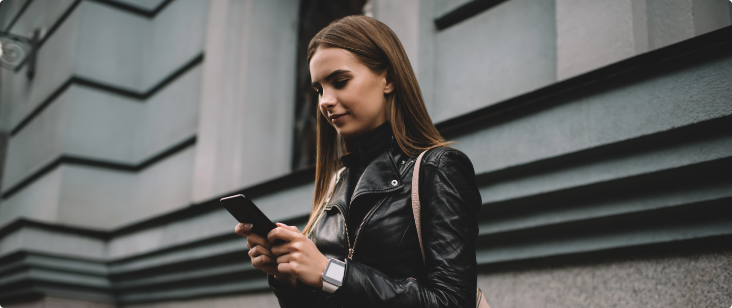Mulher usando celular na rua Fotografia de mulher de jaqueta de couro preta, com cabelo solto e alça de bolsa bege, segurando um celular com as duas mãos, perto de um prédio.