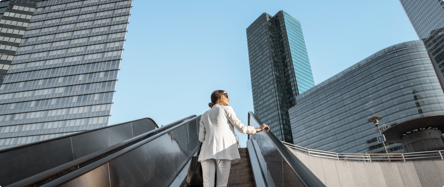 Empresária C6 Conta Global Empresas Mulher de cabelos presos e óculos escuros, vestindo blazer e calça brancos, sobe uma escada rolante ao ar livre. Ela carrega uma bolsa preta no ombro e olha para cima, com edifícios altos e modernos ao fundo. O céu está claro e sem nuvens, sugerindo um ambiente urbano corporativo.