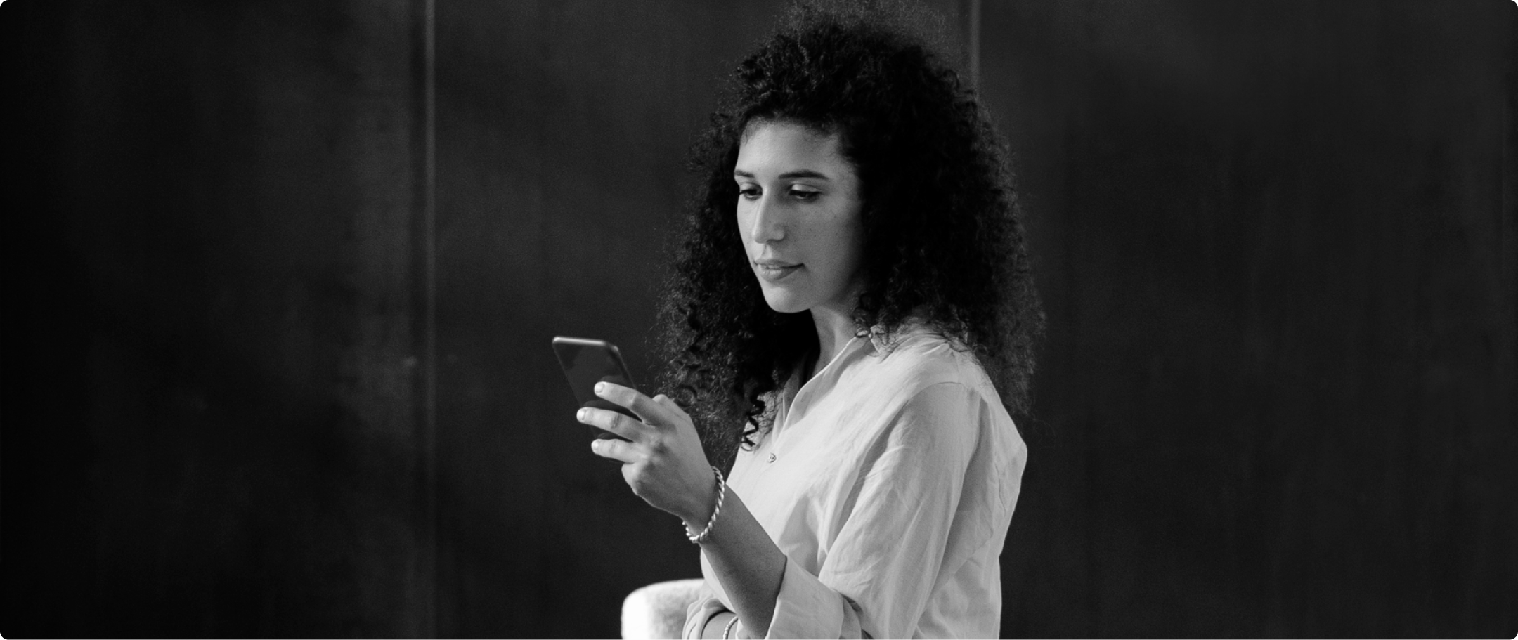 Fotografia em preto e branco de mulher branca com cabelo preso e blusa branca, segurando um celular com a mão esquerda