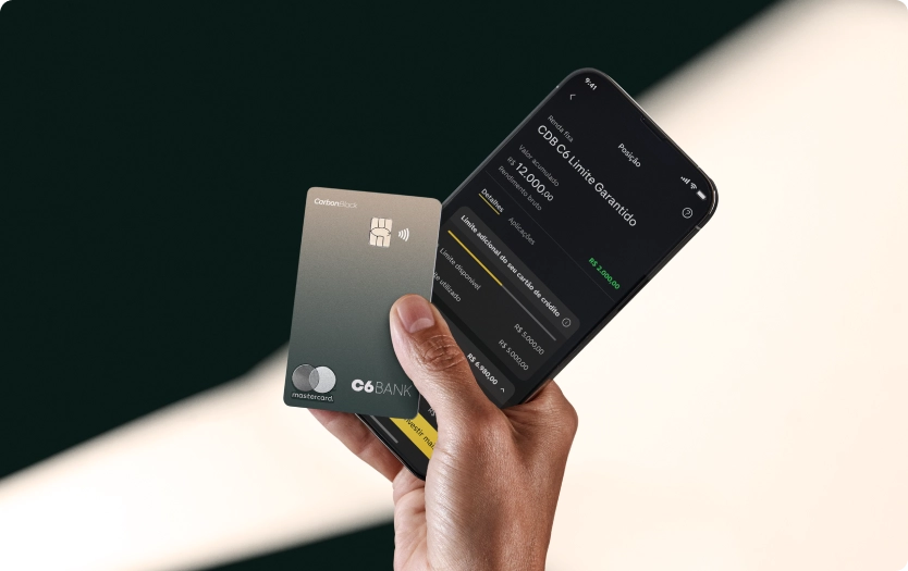 Mão segura um Cartão C6 Carbon Mastercard Black Boreal Forest e um celular com o app C6 Bank.