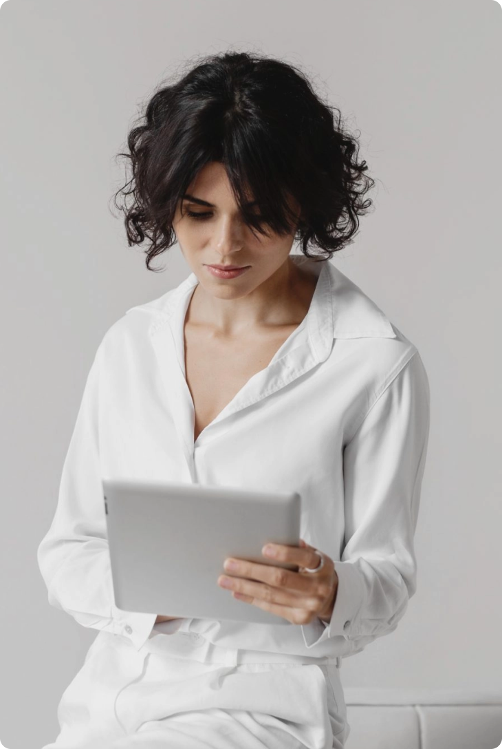 Mulher de camisa social branco, com cabelo curto ondulado preto, encostada em um sofá branco, segurando um tablet.