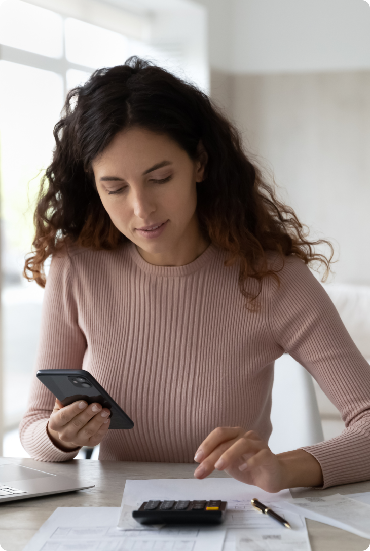 Mulher sentada em uma mesa, fazendo cálculos na calculadora e segurando o celular em outra mão.