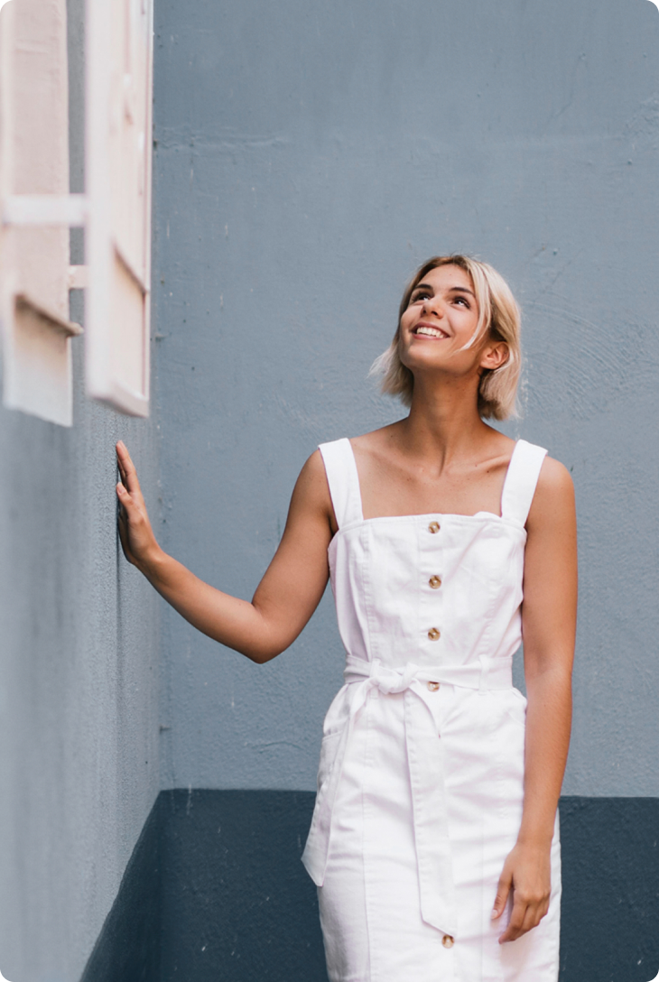 Fotografia de mulher com vestido branco e cabelo loiro, apoiada em uma parede azulada