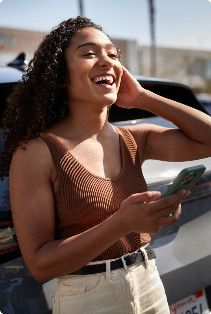 Mulher sorridente ao ar livre segura um celular e está ao lado de um carro, com fundo urbano desfocado.