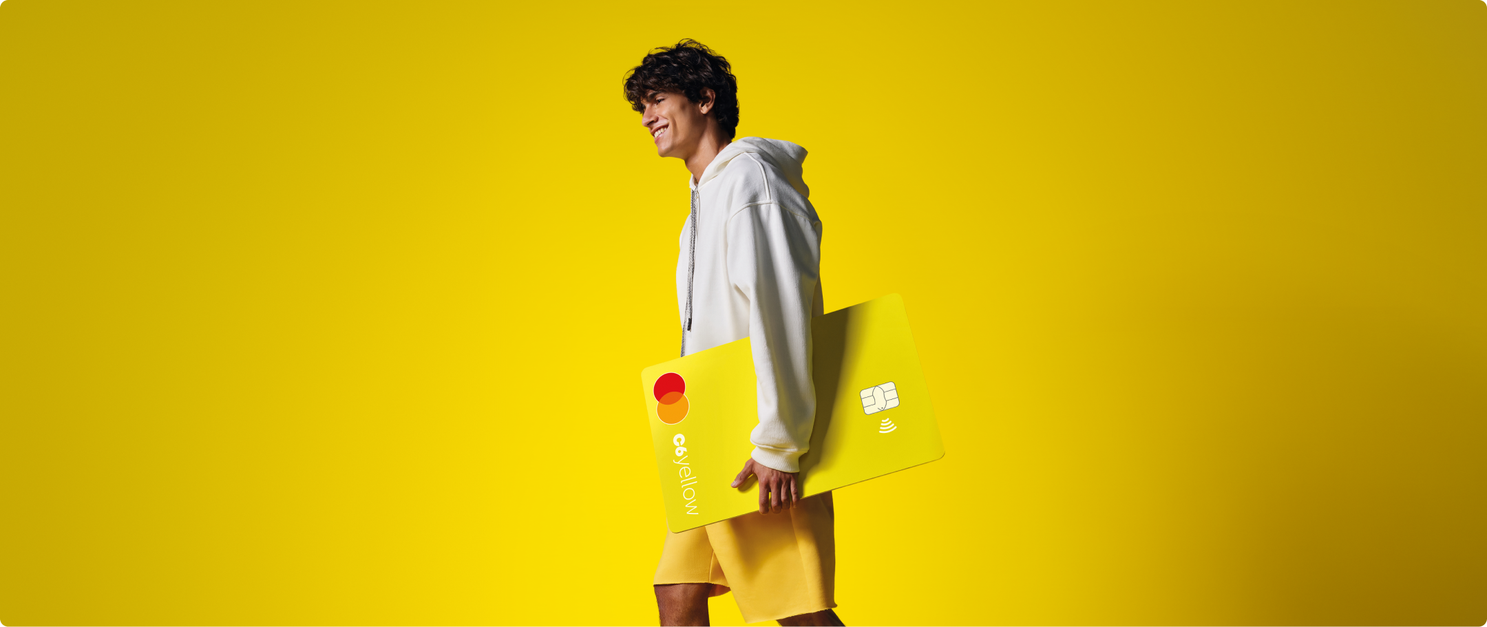 Menino com cartão C6 Yellow SHOOTING 25-26 Menino sorri enquanto caminha segurando cartão C6 Yellow ampliado. Fundo e roupas seguem tom amarelo vibrante.