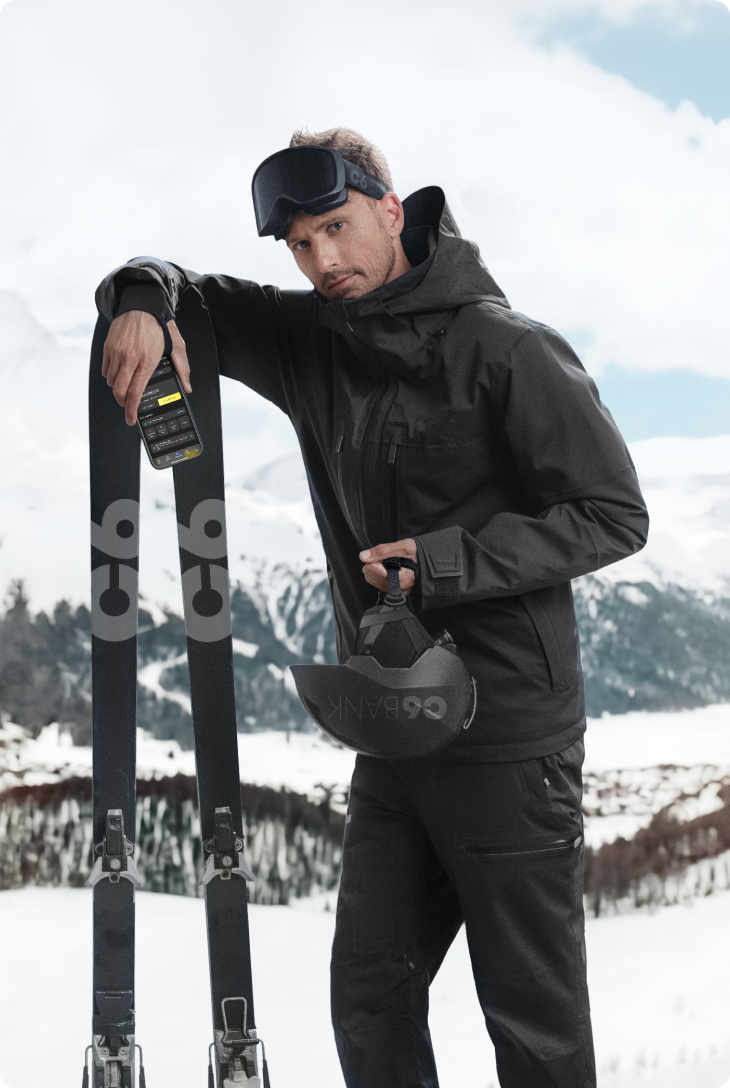 Homem em paisagem de neve com montanhas ao fundo, usando roupa de inverno, apoiada em esquis, segurando capacete, com celular exibindo app do C6 Bank, posicionado sobre o equipamento.