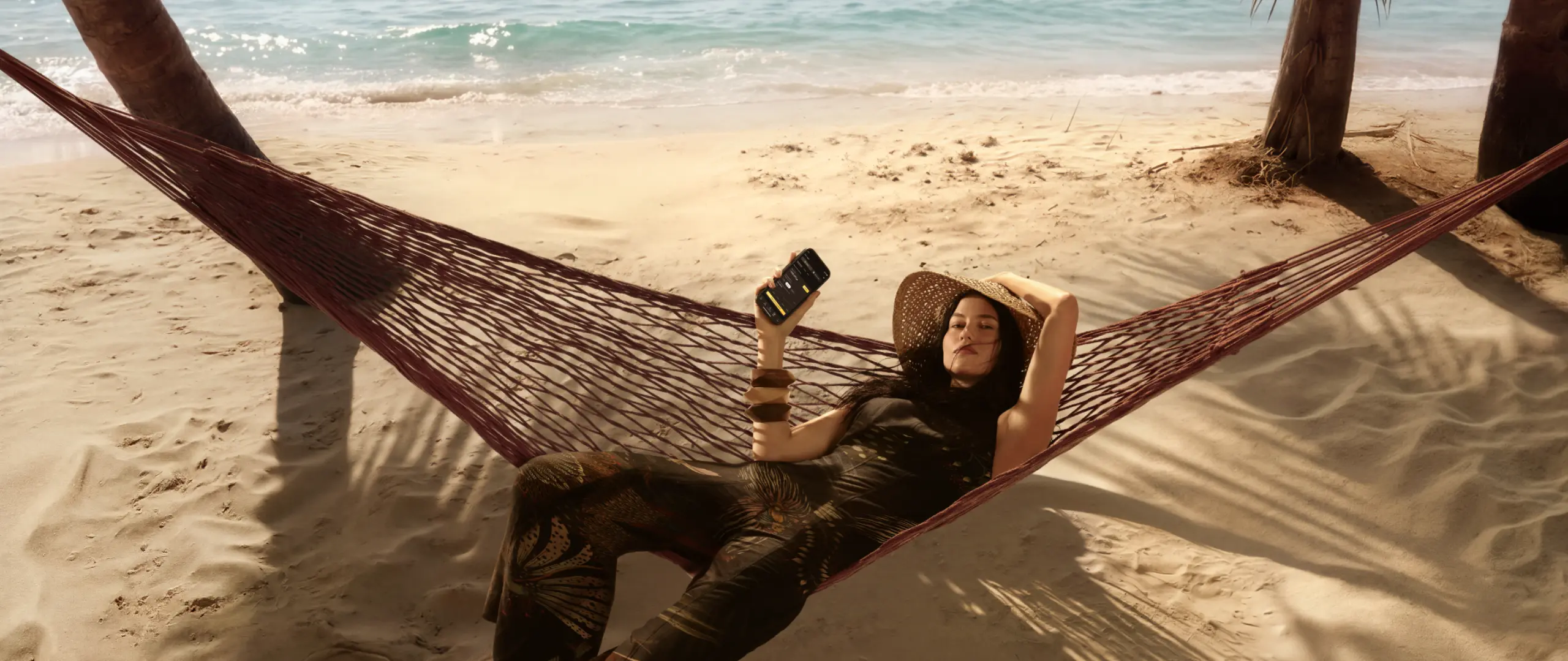 Mulher deitada em rede na praia S 26-27 Mulher deitada em rede sobre a areia com mar ao fundo, usando chapéu e roupa estampada, segurando celular com um braço apoiado atrás da cabeça.