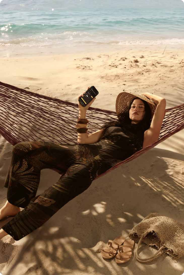 Mulher deitada em rede sobre a areia com mar ao fundo, usando chapéu e roupa estampada, segurando celular com um braço apoiado atrás da cabeça.