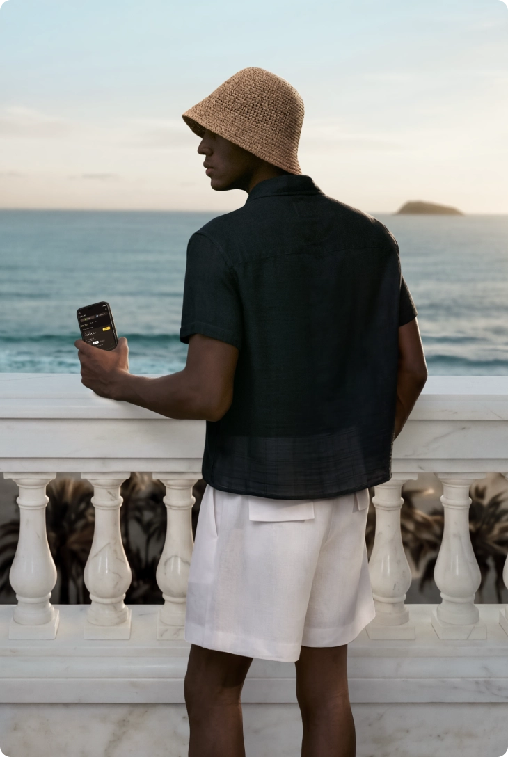 Homem em pé em varanda com vista para o mar, usando chapéu e roupa clara, segurando celular enquanto observa o horizonte.