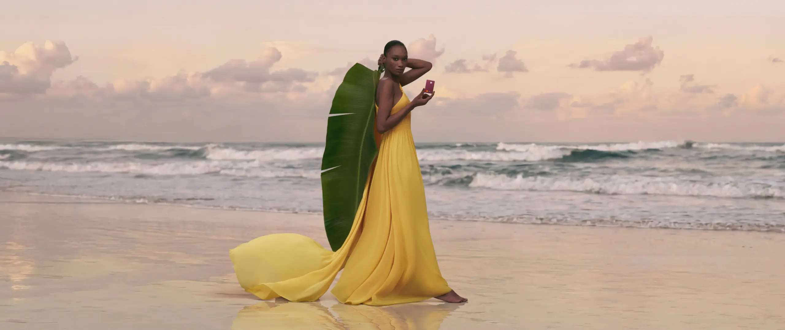 Mulher em praia segurando folha de bananeira S 26-27 Mulher em pé na praia com mar ao fundo, usando vestido amarelo e segurando cartão C6 Conta Global na cor vinho, com grande folha verde apoiada ao lado do corpo.