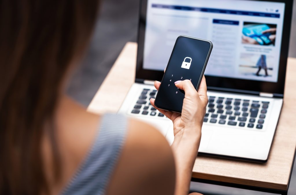 Mulher segura celular com cadeado na tela em frente ao computador